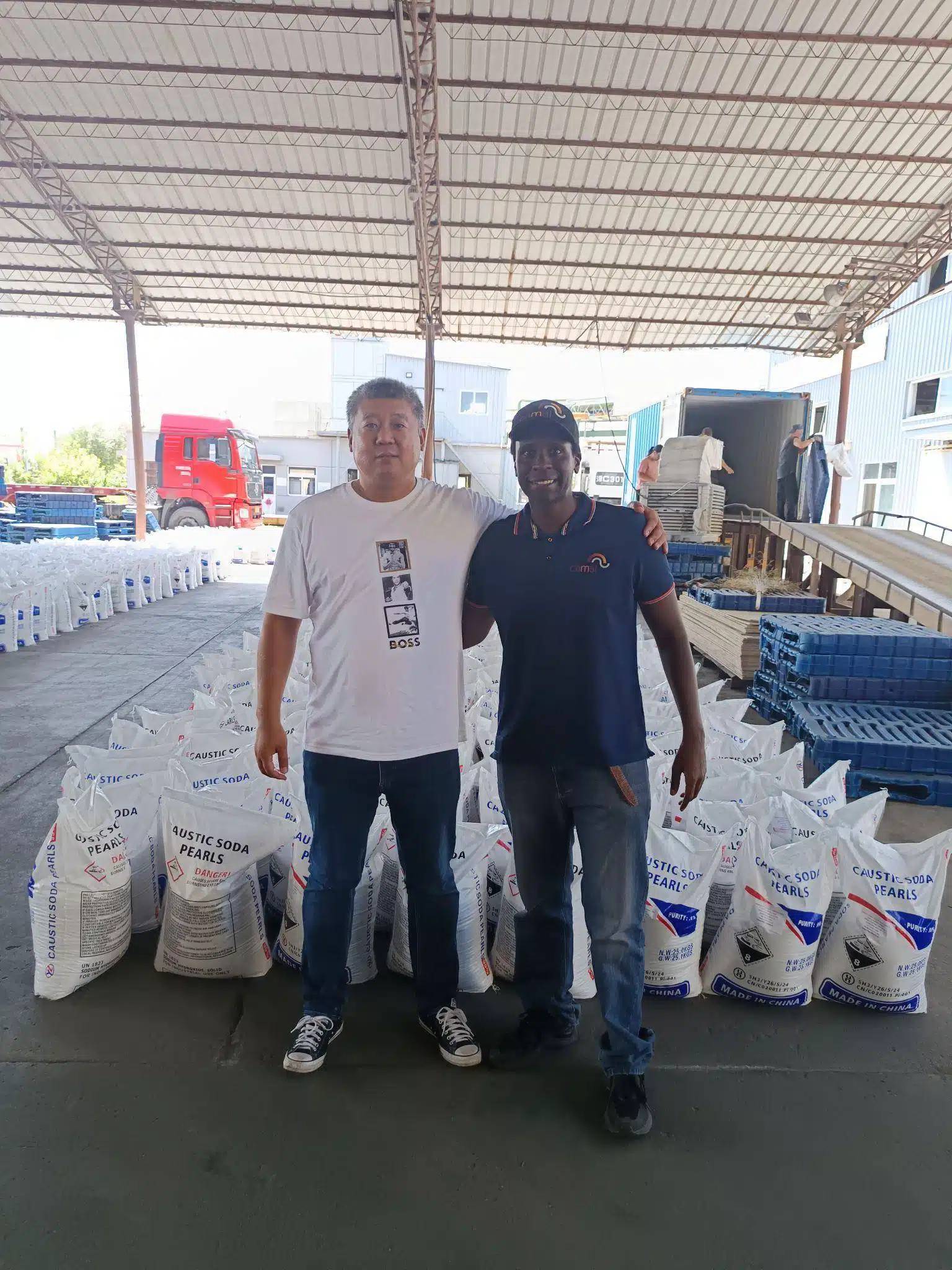 Two men standing inside a warehouse with stacked 25 kg bags of caustic soda pearls prepared for shipment.